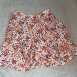 Blommig kjol från Lindex - Säljer en söt blommig kjol från Lindex i storlek XS. Kjolen har ett färgglatt mönster med orange och blå blommor på en vit bakgrund. Den har volanger. Jag älskar kjolen med den har tyvärr blivit för liten.