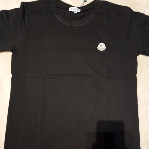Svart t-shirt från Moncler - Snygg svart t-shirt från Moncler med rund halsringning och korta ärmar. Den har en broderad Moncler-logga på bröstet. Perfekt för en stilren look. Liten i storlek (passar som L) 