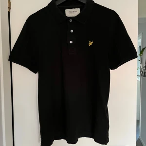 Svart pikétröja från Lyle & Scott - Säljer en stilren svart pikétröja från Lyle & Scott med klassisk krage och knappar framtill. Tröjan har en broderad logotyp i gult på bröstet. Perfekt för en avslappnad men snygg look.