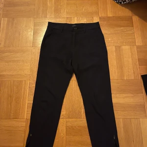 Svarta byxor från Gabba - Snygga svarta byxor från Gabba med diskret rutmönster. Byxorna har en dragkedja vid bensluten och en knapp i midjan. Perfekta för en stilren look.