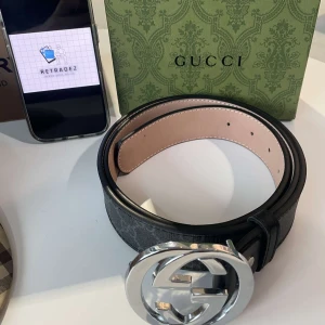 Gucci Bälte - Snyggt svart bälte från Gucci med en stor, rund spänne i silverfärg. Bältet har en klassisk design med Gucci-logotypen på spännet. Perfekt för att ge en lyxig touch till din outfit.