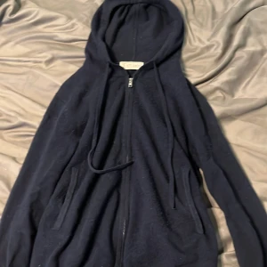 kashmir hoodie från S&T Studios - Säljer en mörkblå kashmir och merinoull hoodie från S&T Studios i storlek XS. Den är tillverkad av 70% merinoull och 30% kashmir, vilket gör den supermjuk och bekväm. Hoodien har dragkedja och justerbar huva med snören. Perfekt för kyliga dagar!