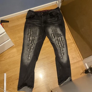 Svarta jeans med amerikanskt flaggmönster från Rock Rebel - Coola svarta jeans från Rock Rebel med ett unikt amerikanskt flaggmönster på framsidan. Jeansen har en edgy design med slitna detaljer och en läcker dödskallelogga på baksidan. Perfekta för dig som vill sticka ut! Storlek: W34L32