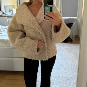 Beige Gina jacka  - Storlek S,  Använd endast fåtal gånger, nyskick  Kort teddyjacka med oversize fit. Teddyjackan är beige och har knäppning fram. Den har mjuk kvalitet, rak ärm, hög krage, sidofickor och sömdetaljer på ärmarna.