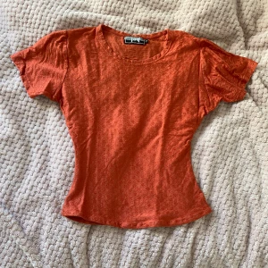 Orange topp med korta ärmar - Säljer en snygg orange topp i storlek M ursprungligen men figursydd så passar XS. Den har korta ärmar och är gjord i ett mjukt material med ett subtilt mönster. Perfekt för både vardag och fest!