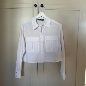 Vit croppad skjorta från Zara - Säljer en vit croppad skjorta från Zara. Skjortan har långa ärmar och två stora fickor framtill. Passar både till vardags och fest!