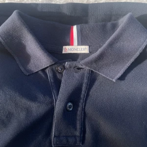 Mörkblå pikétröja från Moncler - Säljer en snygg mörkblå pikétröja från Moncler. Den har en klassisk krage med knappar och Monclers ikoniska logga på ärmen. Perfekt för en stilren och avslappnad look. Tröjan är i bra skick och redo för nya äventyr! Hör av er vid minsta lilla! 