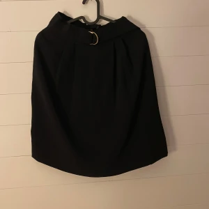 Svart kjol från H&M - Säljer en stilren svart kjol från H&M i storlek 34. Kjolen har en elegant design med ett bälte i midjan och en diskret dragkedja baktill. Perfekt för både vardag och festliga tillfällen!