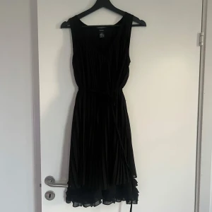 Svart klänning med volangdetaljer - Säljer en elegant svart klänning med volangdetaljer i nederkant. Klänningen är ärmlös och har en normal passform som passar perfekt för festliga tillfällen. Materialet är mjukt och bekvämt, vilket gör den idealisk för både sommar och höst.