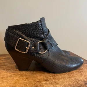 Svarta boots med spänne - Snygga svarta boots i skinn med en cool spetsig tå och klack. De har en bred rem med ett stort metallspänne som ger en edgy look. Perfekta för höst/vår, och de passar både till vardags och fest. Skorna har en dragkedja på sidan. Fin ny sula.