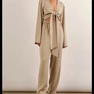 Beige jumpsuit med knytning - Säljer en superfin beige jumpsuit i satin med långärmad design och knytning framtill. Den har en loose passform och är perfekt för fest.