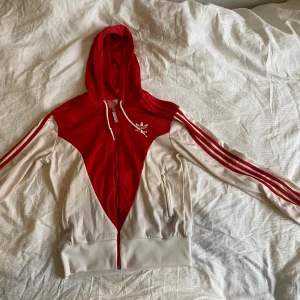 Röd och vit huvtröja från Adidas - Säljer en snygg röd och vit huvtröja från Adidas. Den har en dragkedja framtill och klassiska tre ränder längs ärmarna. Strlk 38 och ganska kort (sitter i midjan) 