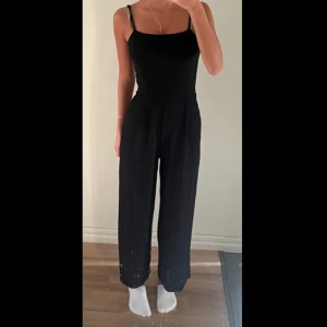 Svart ärmlös jumpsuit - Säljer en stilren och elegant svart jumpsuit. Den är ärmlös med smala axelband och har en tight passform upptill som övergår i vida byxben. Perfekt för både fest och vardag!