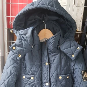 Mörkblå quiltad jacka från Chiara Forthi - Säljer en snygg mörkblå quiltad jacka från Chiara Forthi. Jackan har ett bälte i midjan och guldknappar som ger en lyxig känsla. Den är perfekt för höst och vår, med långa ärmar och flera praktiska fickor. En stilren och elegant jacka för alla tillfällen!