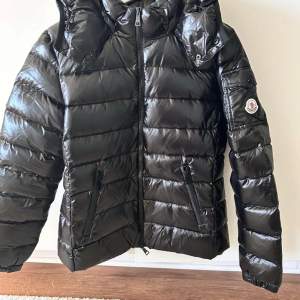 Säljer en snygg och varm svart dunjacka från Moncler. Jackan har en glansig finish och är perfekt för kalla vinterdagar. Den har en dragkedja framtill och en praktisk huva. Moncler-loggan syns på ärmen. QR - kod finns. som äkthetsbevis. 