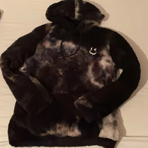 Mönstrad pälshoodie - Säljer en cool och mysig pälshoodie med huva i ett unikt mönster. Hoodin har långa ärmar och en mjuk känsla, perfekt för att hålla värmen. Den har en mörk färgpalett med inslag av ljusare toner.