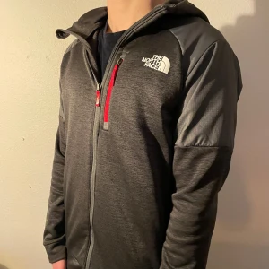Grå flis från The North Face - Säljer en grå/svart flis från The North Face med huva och dragkedja. Flisen har en röd detalj vid bröstfickan och är långärmad. Väldigt skön perfekt passform