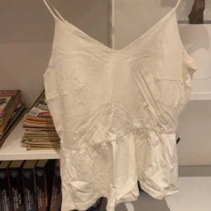 Vit ärmlös topp - Säljer en stilren vit ärmlös topp med smala axelband. Toppen har en enkel design med en lätt rynkad nederdel som ger en avslappnad look. Perfekt för varma dagar eller som ett basplagg i garderoben.