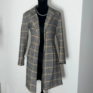 Rutig kappa med bälte - Snygg rutig kappa i svart, vit och gul med klassiskt mönster. Den har långa ärmar och ett bälte i midjan för en elegant passform. Perfekt för att ge din outfit en stilfull touch.