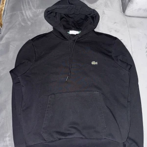 Svart hoodie från Lacoste - Säljer en stilren svart hoodie från Lacoste med klassisk passform. Tröjan har en känguruficka framtill och en justerbar huva med dragsko. Perfekt för en avslappnad look. Den är i fint skick och väll skött ny pris på denna kofta liger runt 1,700kr mitt pris endast 1000kr 
