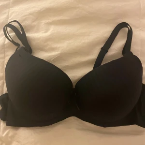 Svart bh med rosett - Säljer en snygg svart pushup bh med en liten rosett mellan kuporna. Bh:n har justerbara axelband och en slät yta som ger en elegant look. 