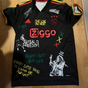 Svart fotbollströja med Bob Marley-tema - Säljer en unik svart fotbollströja från Adidas med Bob Marley-tema. Tröjan har färgglada detaljer i rött, gult och grönt samt tryck med texter som 'One Love One Heart' och 'Football is Freedom'. Ziggo-loggan pryder framsidan och Bob Marleys bild finns på baksidan med texten 'Live the Life You Love'.