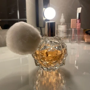 Parfym med fluffig boll - En elegant parfymflaska med ett unikt geometriskt mönster och en fluffig vit boll som dekoration. Flaskan är fylld med en gyllene doftvätska och har en silverfärgad kork. Perfekt för den som vill ha en stilfull och lekfull detalj på sminkbordet.