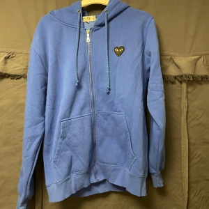 Cdg hoodie  - Säljer en blå hoodie från Play med en unik hjärtdetalj med ögon på bröstet och ryggen. Tröjan har dragkedja och en bekväm passform med långa ärmar och fickor framtill. Perfekt för en avslappnad stil.