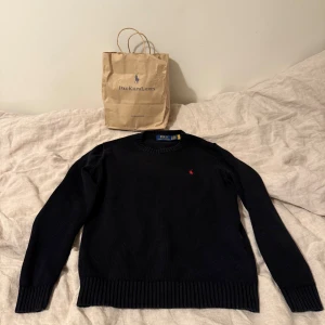 Svart stickad tröja från Ralph Lauren - Säljer en stilren svart stickad tröja från Ralph Lauren med det klassiska röda logotypbroderiet på bröstet. Tröjan har långa ärmar och ribbstickade muddar vid ärmslut och nederkant. Perfekt för en elegant och avslappnad look. Tröjan är i fint skick. Kom privat för frågor och funderingar 😎👍🏻