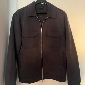 Svart overshirt  från Elvine - Snygg svart overshirt från Elvine med dragkedja framtill och två bröstfickor med lock. Jackan har en klassisk krage och långa ärmar, perfekt för en stilren look. Mycket bra skick. Nypris 2299kr. Hör av dig vid frågor.