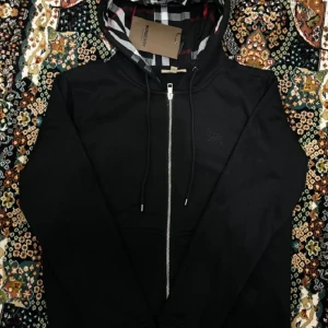 Svart Burberry Hoodie  - Säljer en stilren svart hoodie från Burberry med dragkedja och klassiskt rutigt mönster i fodret. Perfekt för en casual look med en touch av lyx. Huvan har snörning för justerbar passform. Hör av er för de minsta lilla