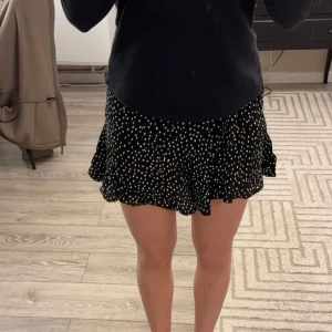 Svart kortkjol med vita prickar och inbyggda shorts - Snygg svart kortkjol med vita prickar och volangdetaljer. Kjolen har en omlott-design som du kan knyta. Kjolen har även inbyggda shorts i sig. Mycket bra skick storlek s