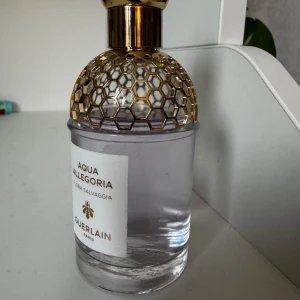 Aqua Allegoria Flora Salvaggia från Guerlain - Elegant parfymflaska från Guerlain, Aqua Allegoria Flora Salvaggia. Flaskan har en stilren design med ett gyllene bikakemönster på toppen och en transparent kropp som visar den ljusa vätskan inuti. Perfekt för den som älskar en fräsch och blommig doft. Väldigt lite använd ❤️