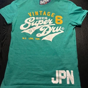 Grön t-shirt från Superdry - Snygg grön t-shirt från Superdry med vintage-tryck i vitt och gult. T-shirten har korta ärmar och en klassisk passform. Tröjan är sparsamt använd och i bra skick. Det är bara att höra av sig vid eventuella frågor😁