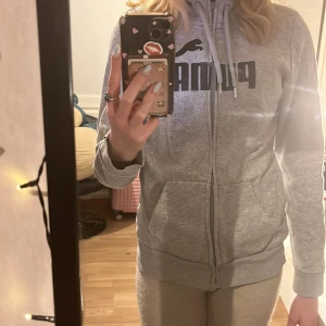 Grå hoodie från Puma - Säljer en grå hoodie från Puma med dragkedja och justerbar huva. Tröjan har en stor logga på framsidan och praktiska fickor. Perfekt för en avslappnad stil. Inga slitage, ser helt ny ut. Storlek S. köparen står för frakten :)