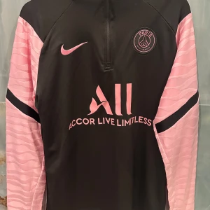 Svart och rosa PSG fotbollströja - Snygg långärmad PSG fotbollströja från Nike i svart med rosa detaljer. Tröjan har en halv dragkedja framtill och klubbens emblem på bröstet. Perfekt för fotbollsfans som vill visa sitt stöd med stil.