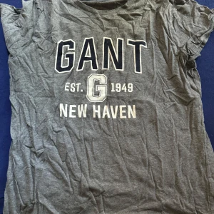 Grå t-shirt från GANT - Säljer en grå t-shirt från GANT med tryck 'GANT EST. 1949 NEW HAVEN' i vitt och mörkblått. T-shirten har en klassisk rund hals och korta ärmar. Perfekt för en avslappnad stil.