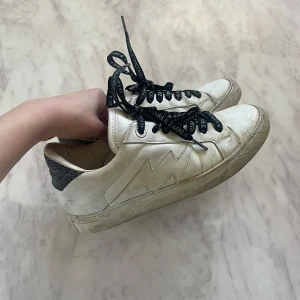 Vita sneakers från Zadig & Voltaire - Snygga vita sneakers från Zadig & Voltaire med svart glittriga snören som har textdetaljer. Skorna har en stilren design med en blixtformad detalj på sidan och en grå hälkappa! De är något missfärgade under skorna samt inuti på sulan, då jag hade dem på flaket, så dem fick lite rost på sig. Dustbagen följer även med!💕✨