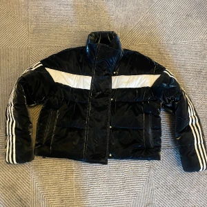 Svart och vit pufferjacka från Adidas - Snygg svart pufferjacka från Adidas med vita detaljer och ikoniska tre ränder på ärmarna. Jackan har en hög krage och dragkedja framtill. Perfekt för kyliga dagar med stil. 🖤