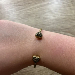Guldarmband med hjärtformade detaljer - Säljer en justerbar gammaldags guld armband med design och hjärtformade ändar dekorerade med små glittriga stenar. Perfekt för att ge en touch av glamour till din outfit.