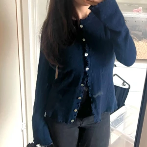 Mörkblå kofta med volangdetaljer - Säljer en mörkblå äkta cashmere kofta med fina volangdetaljer längs knäppningen och vida ärmar. knappar med blommönster. Storlek L men sitter som S. Original pris 2,400