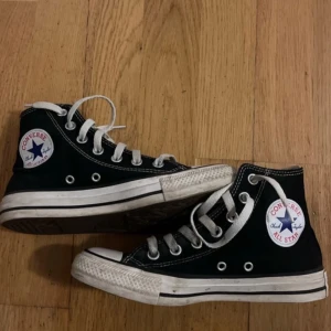 Svarta Converse All Star sneakers - Klassiska svarta Converse All Star sneakers med hög modell och vit snörning. Skorna har den ikoniska logotypen på sidan och en vit sula med svarta ränder. Perfekta för en avslappnad stil. Rekommenderar att tvätta skorna då de är lite smutsiga men det kan enkelt tvättas bort! 