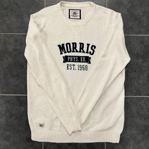 Vit tröja från Morris - Säljer en stilren vit tröja från Morris med tryck 'Morris Phys. Ed. Est. 1968' på bröstet. Tröjan är tillverkad i 100% bomull och har ribbade muddar vid ärmslut och nederkant. Perfekt för en avslappnad stil.