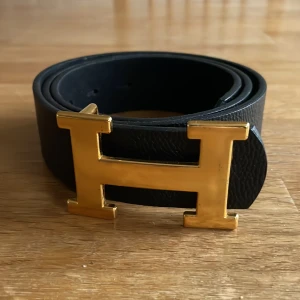 Svart bälte med guldfärgat spänne - Snyggt svart bälte med ett stort guldfärgat H-spänne. Perfekt för att ge en stilren touch till din outfit. Bältet är tillverkat i skinn och har en klassisk design som passar till många olika stilar.