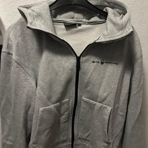 Grå hoodie från Sail Racing - Säljer en grå hoodie från Sail Racing med dragkedja och två fickor framtill. Den har en justerbar huva och logotypen tryckt på bröstet och baksidan av huvan. Perfekt för en avslappnad stil.