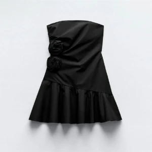 Svart axelbandslös klänning med volang - Elegant svart axelbandslös klänning med volangdetalj i nederkant. Klänningen har dekorativa blommor på framsidan och en smickrande passform. Perfekt för en stilren look.