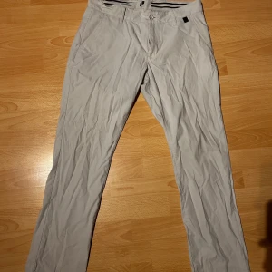 Gråa golfbyxor från Peak Performance - Snygga gråa golfbyxor från Peak Performance med en stilren design. Byxorna har en normal passform. De har knappar och dragkedja framtill samt diskreta bakfickor. Sköna på golfbanan. Inte använda bara skrynkliga.