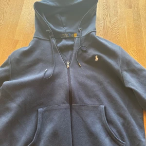 Mörkblå hoodie från Ralph Lauren - Säljer en snygg mörkblå hoodie från Ralph Lauren med dragkedja och klassisk logga på bröstet. Den har en justerbar huva och praktiska fickor framtill. Perfekt för en avslappnad stil. 🧢