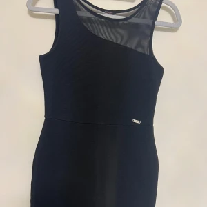 Svart klänning från Guess - Elegant svart klänning från Orsay med en stilren design. Klänningen är ärmlös och har en transparent detalj vid axeln som ger en modern touch. Perfekt för en kväll ute eller en speciell tillställning.