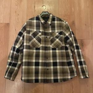 Rutig skjorta från Peak Performance - Snygg rutig skjorta från Peak Performance i beige, svart och grått. Skjortan har långa ärmar och knappar framtill samt på ärmsluten. Perfekt för en stilren look med en touch av outdoor-vibe.